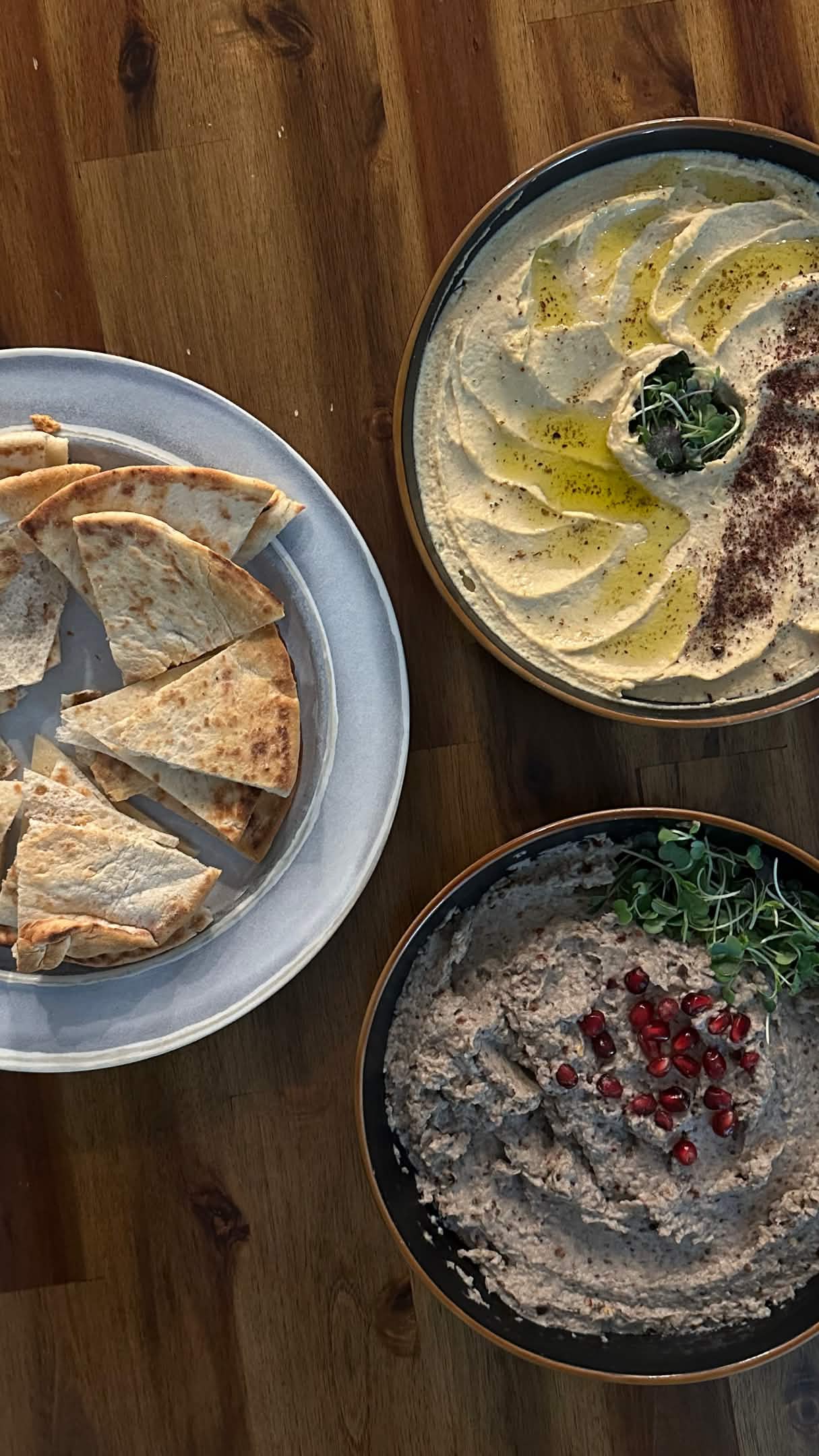 Pita bread to the left, hummus dressed with olive oil, sumac and pea sprouts on the top right, and baba ganoush with pomegranate seeds on the bottom right