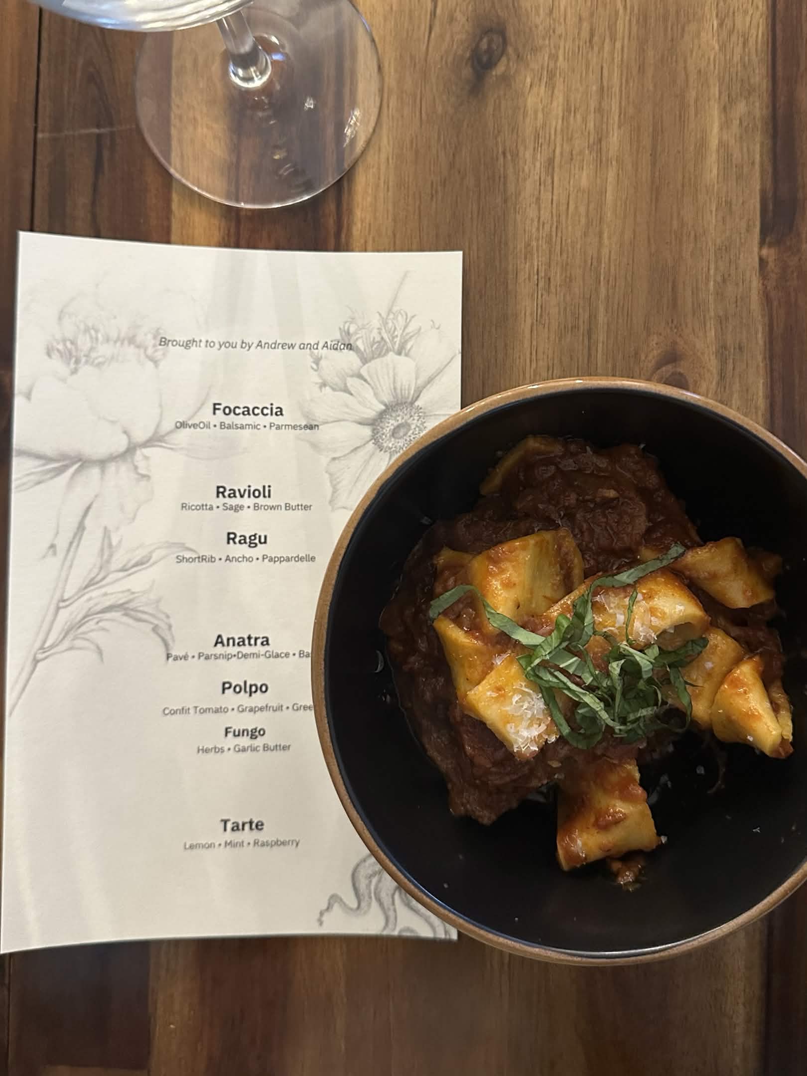 Short rib ragu and papperdelle, garnished with tarragon, plated atop dinner menu with wine glass in the top left