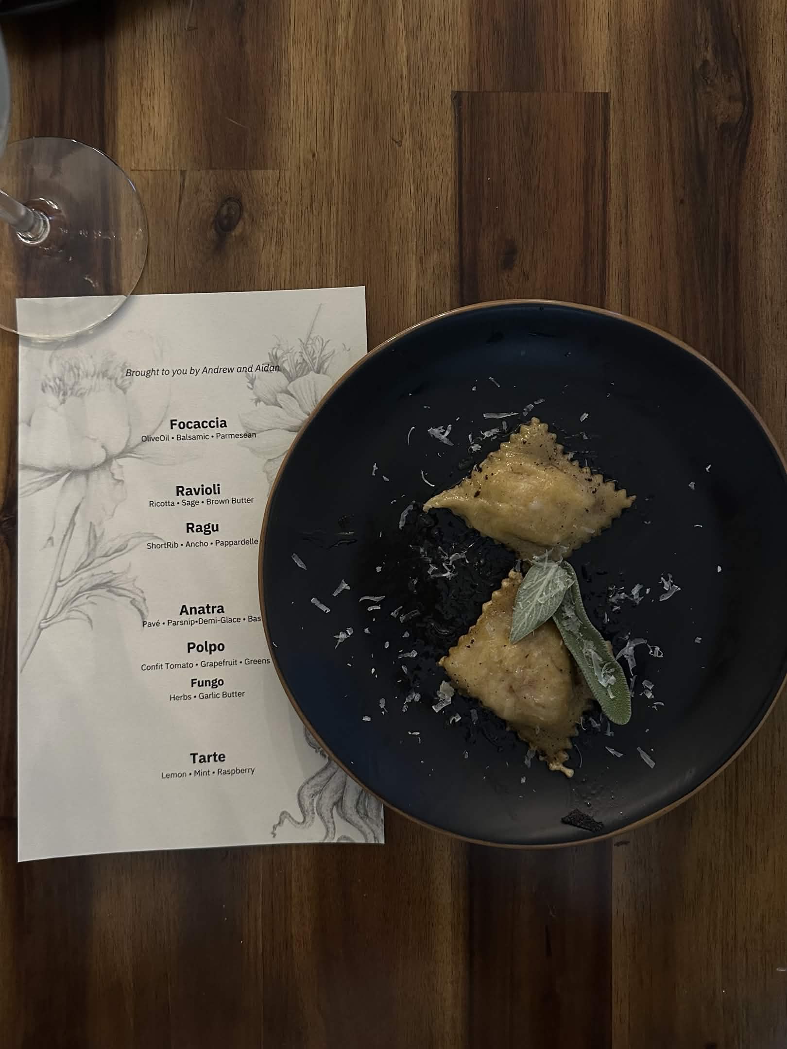 Ravioli in sage brown butter sauce, garnished with fresh sage and grated parmesan, plated atop dinner menu with wine glass in the top left