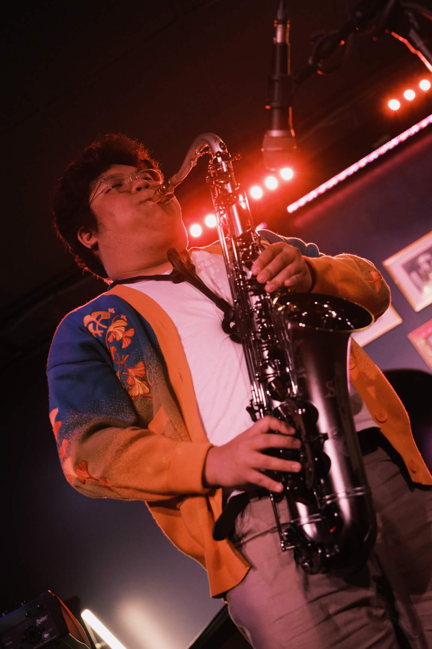 Playing sax into a microphone in gold and green ginko cardigan, backlit by red overhead lights
