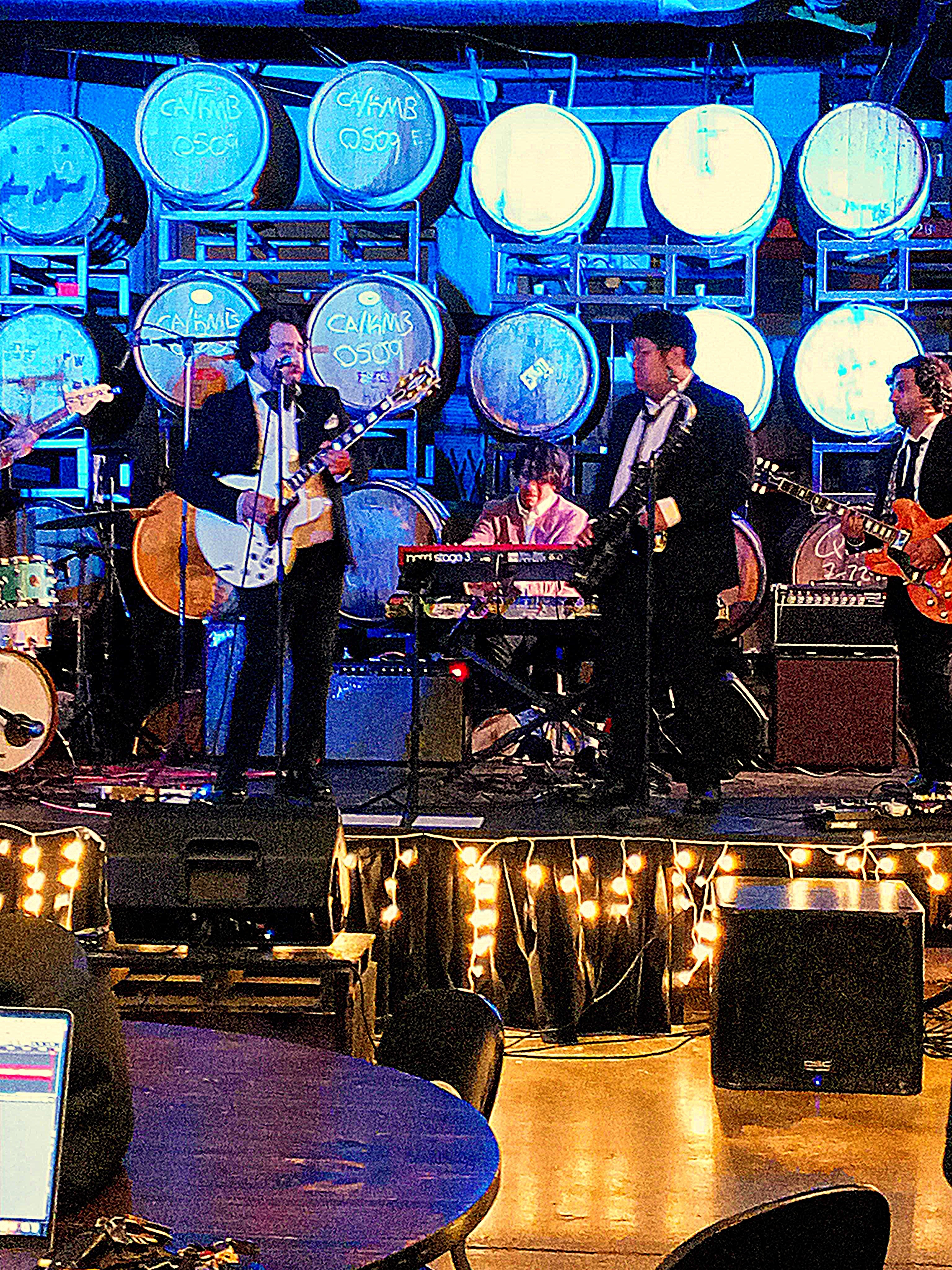 Grainy photo of stage show on raised platform backed with beer kegs. Band is wearing black suits