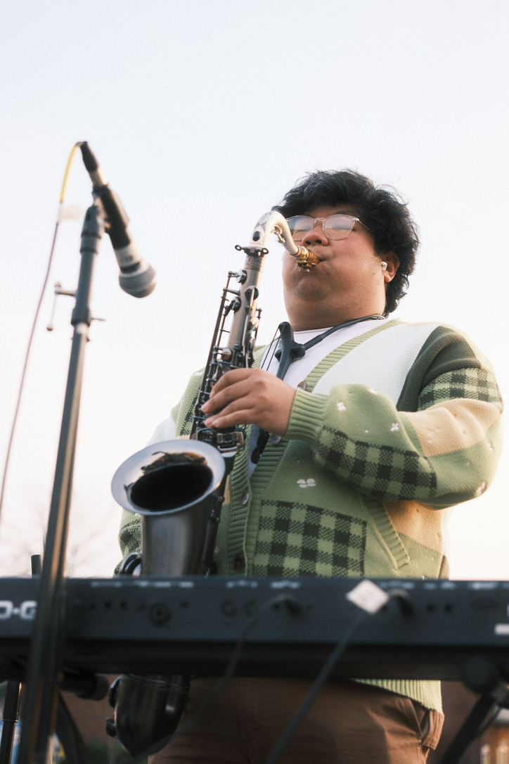 Playing sax into a microphone, wearing a green and white checkered cardigan